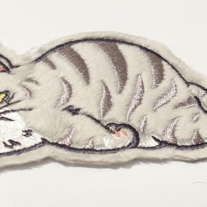 Puede incluir: Parche bordado con un gato gruñón en tonos grises y blancos. El gato está tumbado con la cola enroscada y tiene ojos amarillos y bigotes negros. El parche tiene una textura suave y un contorno cosido.