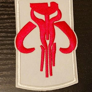 May include: Embroidered patch with a red, stylized symbol on a white background. The symbol has curved, wing-like shapes and vertical lines, all outlined with a white border. The patch is rectangular with rounded corners.