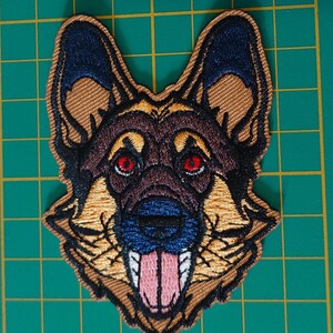 May include: An embroidered patch featuring the head of a German Shepherd dog. The design showcases tan and black fur, pointed ears, bright red eyes, a dark blue nose, and a pink tongue hanging out, all outlined with a thick tan border.