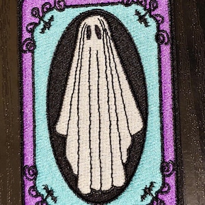 May include: An embroidered iron-on patch featuring a white ghost with a purple and blue frame. The frame has a decorative swirl design.