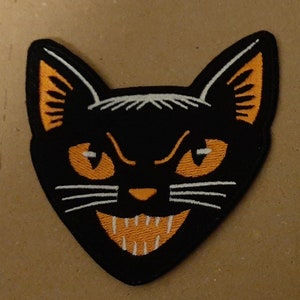 May include: Black cat face embroidered patch with orange eyes and a white mouth. The patch is on a brown background.