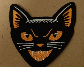 Op de afbeelding: Zwarte kattenkop geborduurde patch met oranje ogen en een witte mond. De patch is op een bruine achtergrond.