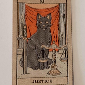May include: A tarot card featuring a black cat with yellow eyes, seated in front of a red curtain. The card is labeled "JUSTICE" and includes a scale and sword. The card has a beige border and the Roman numeral "XI" at the top.