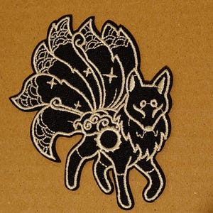 May include: Black and white embroidered patch of a nine-tailed fox with a crescent moon in its tail. The fox is standing on all fours with its tail curled up behind it.