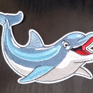 May include: A blue and white embroidered patch of a smiling dolphin with an open mouth. The dolphin is facing right and has a white outline.
