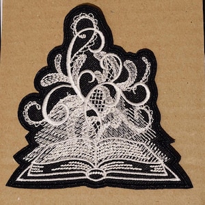 May include: White embroidered patch of an open book with a swirling floral design on a black background.