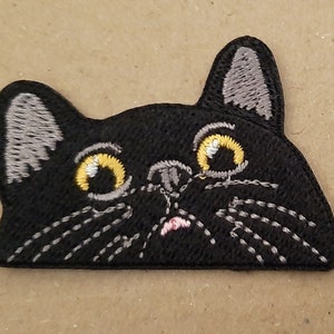 May include: Black embroidered iron-on patch of a cat's face with yellow eyes and a pink nose.