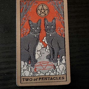 May include: A tarot card depicting two grey cats with collars, set against an orange backdrop with a pentacle symbol. The card reads "TWO of PENTACLES" in a serif font, with a beige border.
