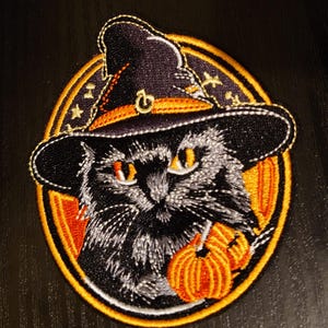 May include: Embroidered patch featuring a black cat wearing a witch's hat and holding a pumpkin. The patch has a yellow, orange, and white border with stars.
