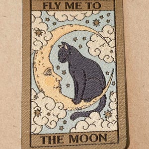 Puede incluir: Tarjeta rectangular con borde marrón que presenta un gato negro sentado en una luna creciente. La luna tiene una cara y está rodeada de nubes y estrellas. El texto "FLY ME TO" y "THE MOON" está arriba y abajo.