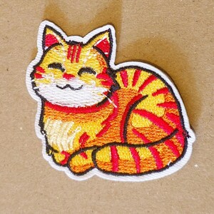 Op de afbeelding: Een geborduurde patch met een gestileerde oranje cyperse kat met donkerrode strepen, een witte snuit en gesloten ogen. De kat is afgebeeld in een opgerolde rusthouding met een witte rand die de gehele vorm van het ontwerp omlijnt.