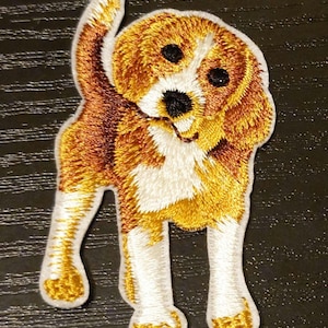 Puede incluir: Parche bordado con un perro beagle de pie. El parche tiene una combinación de colores blanco, marrón y dorado. Los ojos y la nariz del perro son negros. El parche es probablemente para ropa o accesorios.