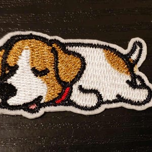 Może przedstawiać: Naszywka haftowana przedstawiająca śpiącego białego i brązowego psa. Pies ma czarne oczy i czerwony język.
