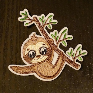 Puede incluir: Parche de planchado bordado de un perezoso marrón con ojos grandes colgado de una rama de árbol verde.