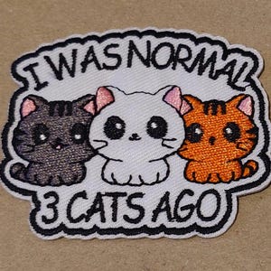 May include: Embroidered patch featuring three cartoon cats in black, white, and orange. The patch has the text "I WAS NORMAL 3 CATS AGO" in a black outline on a white background. The cats have pink ears and large eyes.
