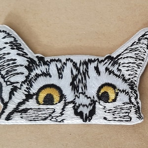 May include: Embroidered iron-on patch of a white cat's face with black stripes and yellow eyes. The cat is looking directly at the viewer.