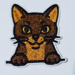 Puede incluir: Parche bordado con un gato de dibujos animados con pelaje marrón y canela, ojos amarillos y bigotes negros. El gato sonríe y parece estar mirando por encima de un borde blanco. El parche está diseñado para su aplicación en ropa o accesorios.