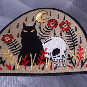 May include: A black cat and a white skull embroidered on a beige patch with red flowers and gold leaves. The patch is shaped like a crescent moon.
