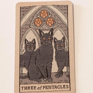 May include: A tarot card featuring three black cats with orange eyes, set against a gothic archway with three pentacles. The card is labeled "THREE of PENTACLES" in a serif font, with a beige border.