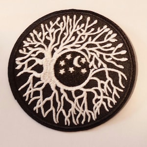 Pode incluir: Remendo circular preto com um desenho de árvore bordado em branco. A árvore apresenta uma lua crescente e estrelas dentro de seus galhos. As raízes se estendem para fora, criando um padrão detalhado e intrincado.