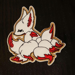 Könnte beinhalten: Weißer gestickter Aufnäher mit einem Kitsune, einem japanischen Fabelwesen, einem Fuchs mit neun Schwänzen. Der Fuchs ist zusammengerollt, seine Schwänze bilden einen Wirbel um seinen Körper. Die Schwänze sind goldfarben umrandet und haben rote Spitzen.