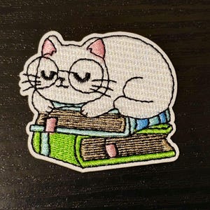 Op de afbeelding: Witte geborduurde patch van een kat met een bril die slaapt op een stapel boeken. De kat is wit met roze oren en een blauw-wit gestreepte pyjama. De boeken zijn groen, bruin en blauw.