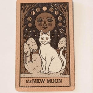May include: A rectangular patch with a white cat, a brown moon with a face, and moon phases. The text "the NEW MOON" is at the bottom. The background features trees and stars, all in shades of brown and beige. A mystical design.