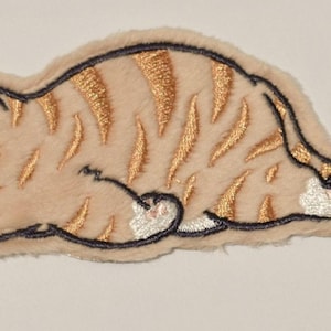 May include: Embroidered patch of a sleeping orange tabby cat with black and gold accents. The cat is lying down with its eyes closed, and its fur has a textured appearance. The patch has a light beige border.