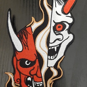 May include: An embroidered patch featuring two Japanese demon masks, one with a red eye and the other with a black eye, both with sharp teeth and horns. The masks are surrounded by flames.
