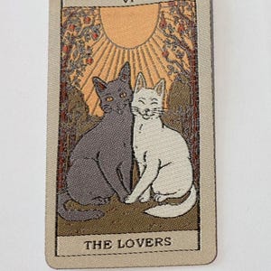 May include: A tarot card illustration showing two cats, a grey one and a white one, side by side. The card has a beige border and features a sun, trees, and the words "THE LOVERS". The card is labelled "VI" at the top.