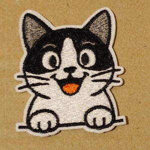 Op de afbeelding: Geborduurde patch met een cartoonkat met een vrolijke uitdrukking. De kat heeft zwart-witte vacht, grote ogen en een oranje tong. De patch is zwart omlijnd en heeft een witte achtergrond. De kat lacht en lijkt een horizontale lijn vast te houden.