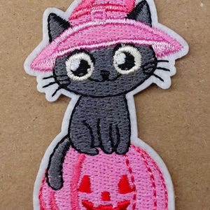 Op de afbeelding: Een geborduurde patch met een donkergrijze kat met grote witte ogen die een felroze puntmuts draagt. De kat zit bovenop een roze pompoen met een uitgesneden jack-o'-lantern gezicht in een donkerdere roze tint, omlijnd in het wit.