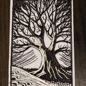 May include: Black and white embroidered patch of a tree with bare branches. The tree is on a white background with a white embroidered ground.