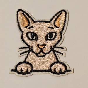 May include: Embroidered patch featuring a stylized cat with a light tan body, black outlines, and tan ears. The cat has large, oval eyes and a small nose. The patch is designed to look like the cat is peeking over a ledge.