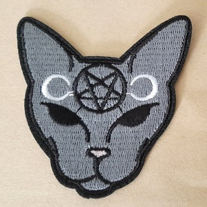 May include: Grey embroidered patch of a cat's face with a pentagram and two crescent moons above its eyes.