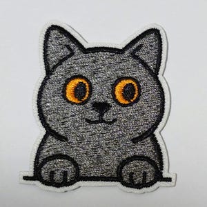 May include: Embroidered patch of a grey cat with large orange eyes, a black nose and mouth, and a white border. The cat is smiling. Suitable for clothing or accessories.