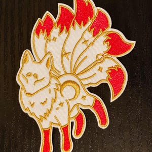 May include: A white embroidered patch depicting a nine-tailed fox with red tails. The fox is outlined in gold thread and has a crescent moon and stars on its body.