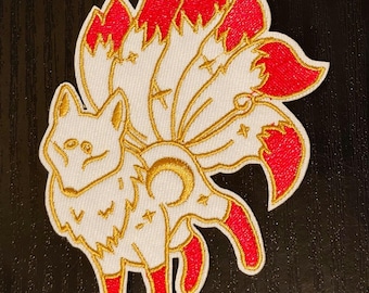 May include: A white embroidered patch of a nine-tailed fox with red tails. The fox is outlined in gold thread and has a crescent moon and stars on its body.