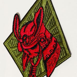 May include: Red and black embroidered patch of a samurai warrior with a helmet and a green diamond-shaped background.