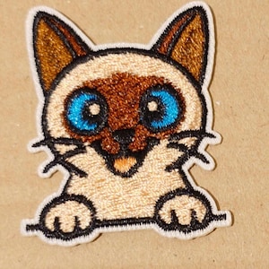 Puede incluir: Parche bordado con un gato siamés con orejas y marcas faciales marrones, ojos azul brillante y una expresión sonriente. El parche tiene un borde blanco y está diseñado para coser o planchar sobre tela. Se ven las patas del gato.