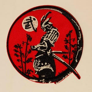 May include: A red circular patch with a black silhouette of a samurai warrior standing in front of a white moon with the Japanese character "武" (pronounced "bu", meaning "martial") on it. The samurai is holding a sword and is surrounded by black grass.