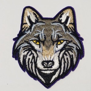 May include: Embroidered wolf patch featuring a detailed design. The wolf's head is depicted with brown, gray, and white thread, with yellow eyes and a purple border. This patch is suitable for jackets, bags, or other fabric items.