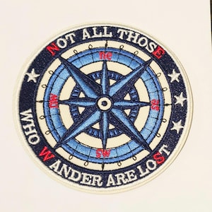 Puede incluir: Parche bordado con un diseño de brújula en tonos azules y blancos, con detalles en rojo. El parche lleva la frase "NOT ALL THOSE WHO WANDER ARE LOST" en un patrón circular alrededor de la brújula. Estrellas blancas en el borde exterior.
