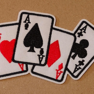 Pode incluir: Patches bordados de quatro ases de cartas de jogar: copas, espadas, ouros e paus. Cada carta apresenta um fundo branco com uma borda preta e o símbolo do naipe em vermelho ou preto. A letra "A" é visível em cada carta.