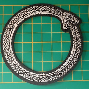 May include: A circular embroidered patch featuring a silver snake biting its own tail, forming an Ouroboros symbol. The snake has a detailed scale pattern in silver thread against a black background, with a black border outlining the entire shape.