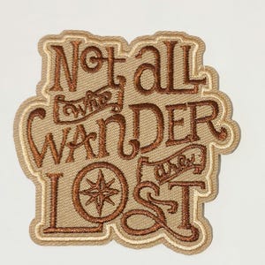 Puede incluir: Parche bordado con el texto "Not all who wander are lost" en hilo marrón sobre fondo beige. El parche presenta un diseño de rosa de los vientos y un borde beige claro.