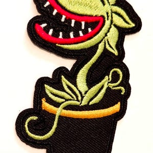 Op de afbeelding: Geborduurde patch met een groene, lachende plant met een rode mond en witte tanden, in een zwarte pot met een oranje rand. De plant heeft groene bladeren en een zwarte omtrek.