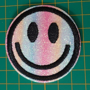 以下が含まれることがあります： スマイリーフェイスのデザインが施された円形の刺繍ワッペン。顔には黒いステッチの輪郭、黒い楕円形の目、黒い曲線状の口があります。ワッペンの背景は、パステル調の虹色のグラデーションで、きらめく虹色の光沢があります。