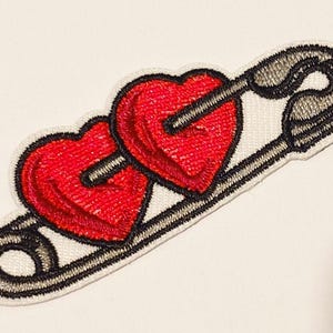 Puede incluir: Parche bordado con dos corazones rojos atravesados por una flecha negra, sobre un fondo blanco. El parche está contorneado con costuras negras, que se asemejan a un imperdible. Ideal para ropa o accesorios.