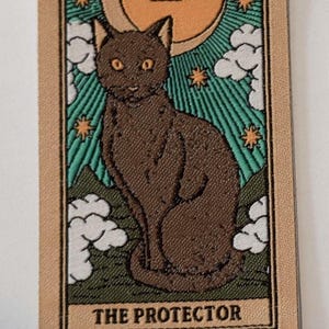 May include: A tarot card featuring a brown cat with yellow eyes, labeled "THE PROTECTOR." The card has a beige border and a background with a crescent moon, stars, and clouds. The Roman numeral "XIII" is at the top.